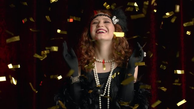 Excited retro styled woman dressed in Roaring twenties era dancing and smiling under confetti rain on dark background. Vintage, party, fashion concept.