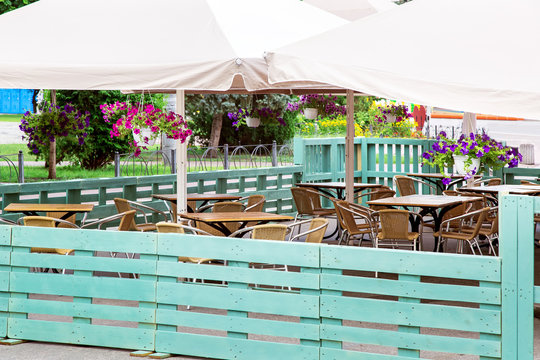 a free wooden table with a wicker chair of an outdoor cafe under an umbrella with hanging flowerpots and blooming petunias fenced with a wooden fence of green painted pallets.