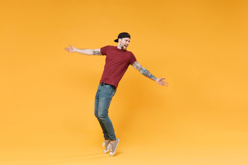 Fototapeta na wymiar Young tattooed man guy in casual t-shirt black cap posing isolated on yellow wall background studio portrait. People lifestyle concept. Mock up copy space. Dancing, standing on toes, spreading hands.