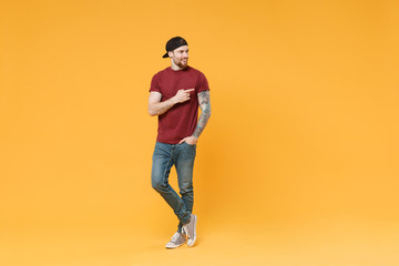 Naklejka premium Smiling young bearded tattooed man guy in casual t-shirt black cap posing isolated on yellow wall background studio portrait. People lifestyle concept. Mock up copy space. Pointing index finger aside.