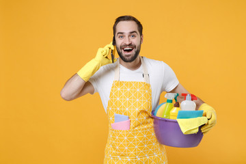 Cheerful young man househusband in apron rubber gloves hold basin with detergent bottles washing...