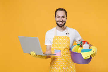 Smiling young man househusband in apron rubber gloves hold basin with detergent bottles washing...