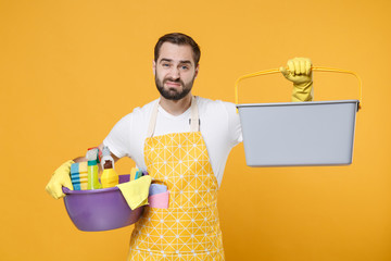 Displeased young man househusband in apron rubber gloves hold basin detergent bottles washing...