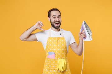 Excited young man househusband in apron hold in hands iron while doing housework isolated on yellow...