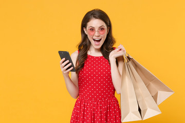 Fototapeta na wymiar Excited young brunette woman girl in red summer dress, eyeglasses posing isolated on yellow background. People lifestyle concept. Hold package bag with purchases after shopping, using mobile phone.