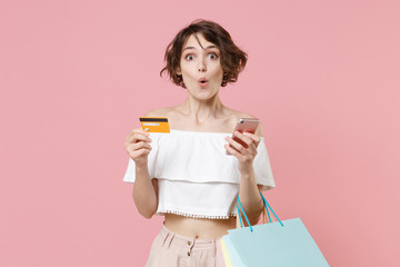 Fototapeta premium Shocked young woman girl in summer clothes isolated on pink wall background. Shopping discount sale concept. Mock up copy space. Using mobile phone, hold package bag with purchases, credit bank card.
