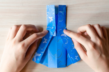 Girl's hands making origami blue dress from butterfly printed paper. Step by step instruction, step16. Mother's day, birhday, summer gift, family leisure time concept. DIY tutorial, flatlay, top view