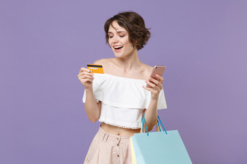 Funny young woman girl in summer clothes isolated on pastel violet background. Shopping discount sale concept. Mock up copy space. Using mobile phone, hold package bag with purchases credit bank card.
