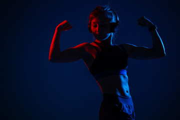 A girl with illuminated relief abdominal muscles shows biceps. Sportswoman in headphones in a neon red and blue light.
