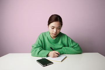 Young asian woman taking down notes in diary wearing casual clothes posing over pink background. Isolated. Free space.