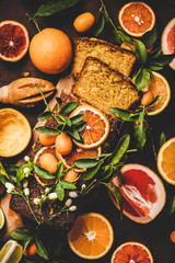 Citrus Mediterranean loaf cake. Flat-lay of almond pound cake with fresh oranges, lemon, kumquats, grapefruits and blossom flowers over dark rusty background, top view. Flourless vegan dessert concept
