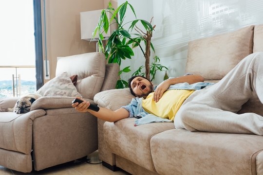 Young Man Holding A TV Remote And Yawning While Resting On A Sofa With His Dog Next To Him