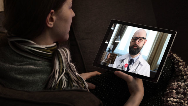 Telemedicine. Consultation with physician through mobile video call. Woman sitting on couch at home and talking to MD on tablet screen.