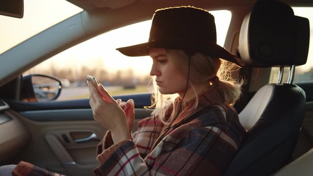 Stunning country girl with blonde hair cowboy hat relaxing on car passenger seat with smartphone using social media application. Portrait. Sunsets.