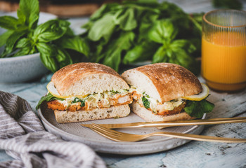 Breakfast table with fresh fried fish sandwich with tartare sauce, lemon and arugula cut in halves, fresh greens and glass of orange juice. Healthy easy breakfast ideas