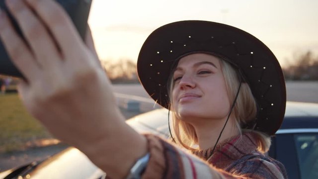 Pretty young western girl posing for polaroid instant camera at sunset. Cheerful blonde cowboy woman taking retro selfie pictures near parked car. Memories concept.