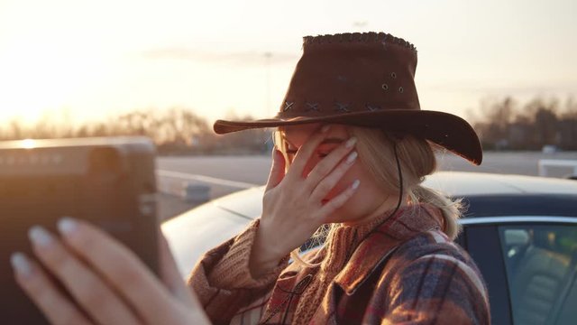 Nice blonde young woman in cowboy hat preparing for selfie pictures fixing her hair and holding instant polaroid camera outdoors at sunset.