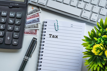 Tax text with notepad, paper clip, keyboard, decorative flower, fountain pen, calculator and banknotes currency on white background. Business and Copy space concept.
