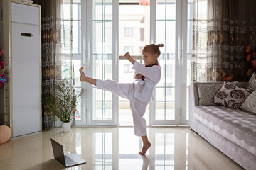 Taekwondo girl in kimono with white belt exercising at home in living room. Online education during coronavirus covid-19 lockdown, self isolation and social distancing concept.