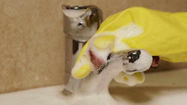 Cleaning. A hand in a protective glove washes a faucet in the bathroom with a washcloth and detergent.
