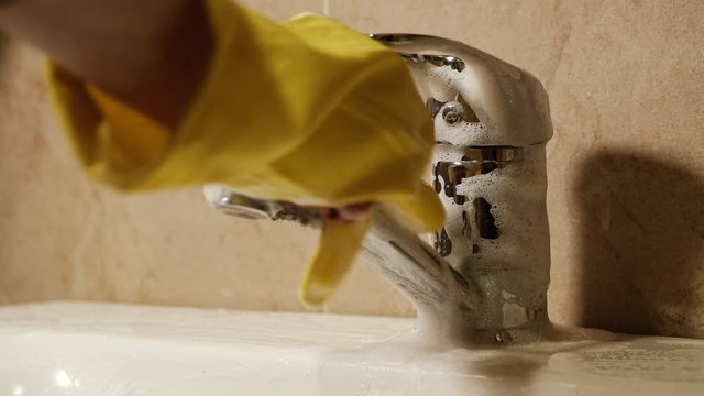 Cleaning. A hand in a protective glove washes a faucet in the bathroom with a washcloth and detergent.
