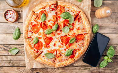 Pepperoni Pizza and smart phone on rustic wooden background. Italian food delivery, call or order online on mobile. Top view.