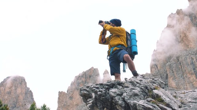 Young man hiker taking pictures on stone mountain with cloudy sky and fog. Yellow jacket, backpack, black beard and beanie. Traveling Dolomites, Italy.
