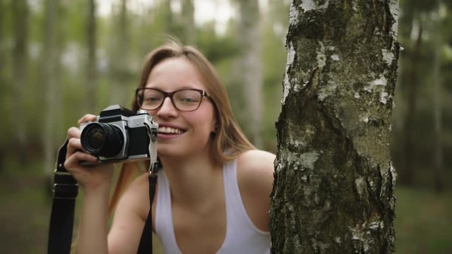 Young caucasian woman hiding behind the tree with a vintage camera. Happy girl photographing the nature.
