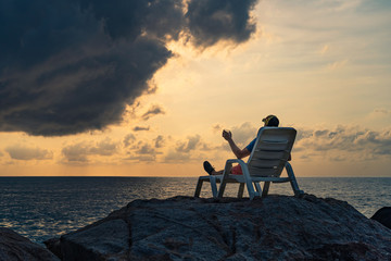 A man with a glass in his hand sits in a lounger on the rocs by the sea against the background of dawn on a summer morning