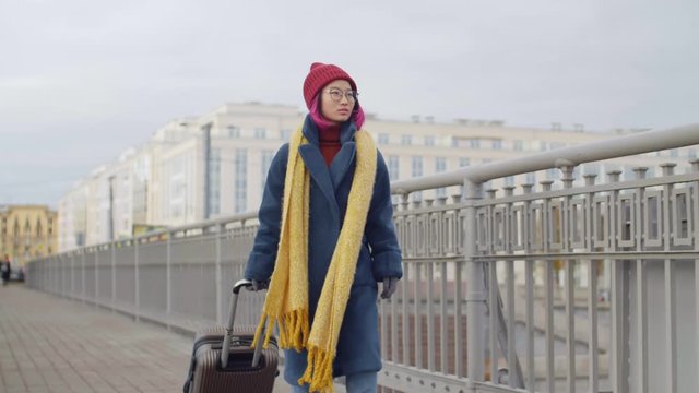 Young Asian female tourist walking with luggage on urban bridge and looking around while searching hotel after arrival in new city on autumn vacation