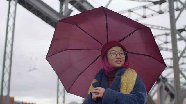 Young beautiful asian woman with pink hair walking with umbrella on urban bridge and smiling while exploring new city on autumn day