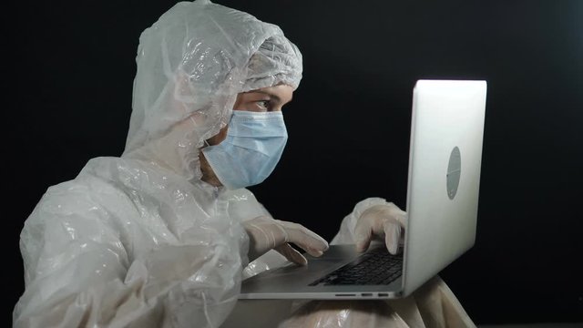 scientist man in a protective suit and mask typing on a laptop at night, develops a vaccine against the pandemic coronavirus covid-19 and writes patient reports in the laboratory conspiracy theory