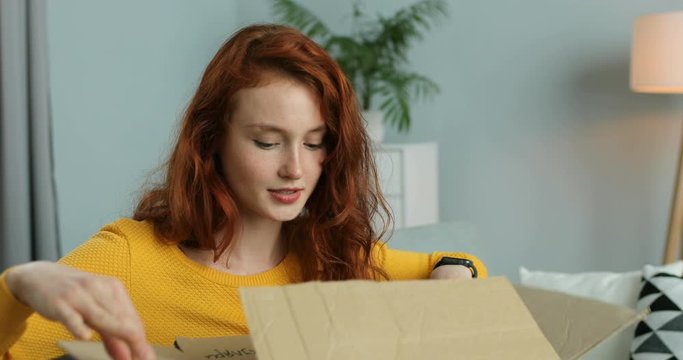 Close up of young Caucasian pretty happy woman sitting on couch in living room and opening carton box. Red-haired attractive cheerful girl getting parcel at home. Mail delivery to house. Lockdown.