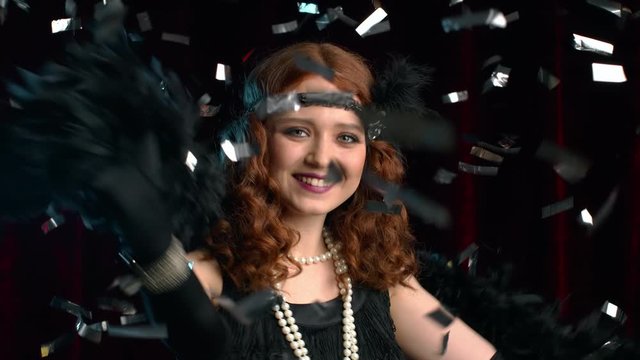 Excited retro styled woman dressed in Roaring twenties era dancing and smiling under confetti rain on dark background. Vintage, party, fashion concept.