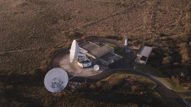 Aerial tracking over a small remote radar station, two dishes and two buildings in a barren moorland setting.