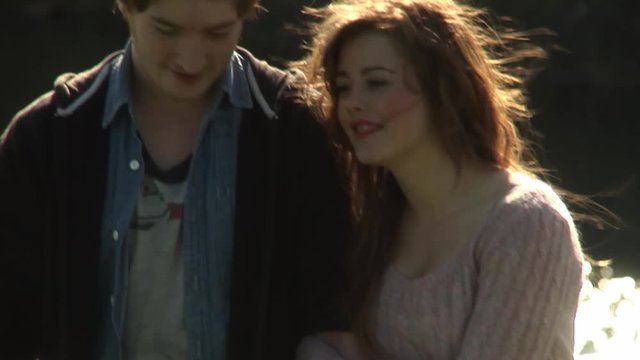 Happy romantic young couple holding hands embraced smiling walking along picturesque lake sunny with warm sunlight glistening off water surface during summers day at the park long shot slow motion