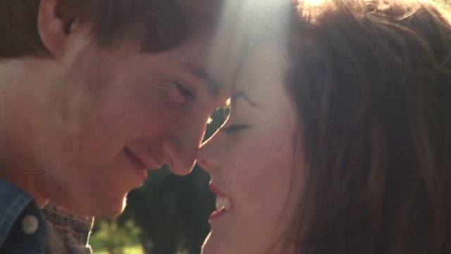 Happy smiling young couple lovingly embrace intimate romantic dreamy moment heads touch looking affectionately into each others eyes on warm sunny day at park slow motion closeup
