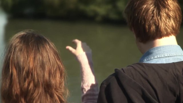 Happy cheerful young couple playfully throwing bread feeding ducks from bridge on warm sunny day at park reverse rear shot back of heads slow motion