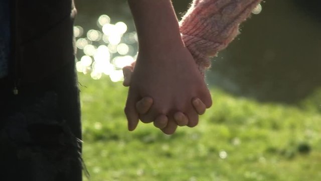Couple holding hands walking along picturesque lake during warm summers day at park lake camera tracking close up shot