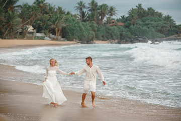 Honeymooners happy couple dressed in white running or jogging on the beach. Groom and bride run along the shore against the wind