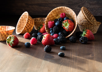 A close up view of waffle bowls with berries spilling out, against a dark background with bright sun shining in.