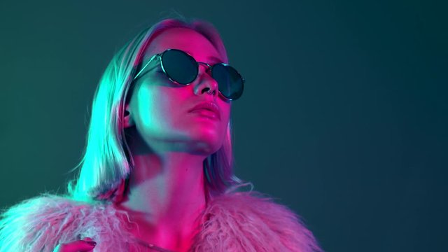 Glamorous hipster teenager in sunglasses and furry coat dancing to music with sunglasses. Portrait of millennial pretty girl with short hairstyle with neon light. Dyed blue and pink hair.