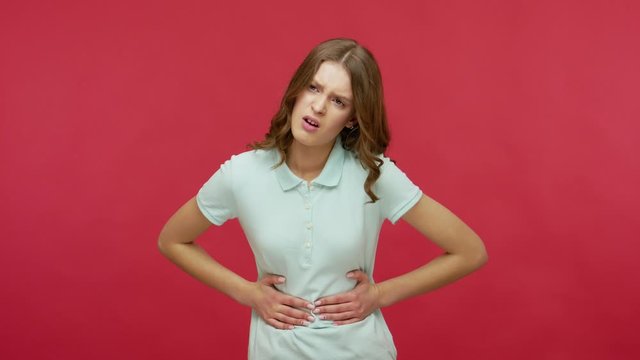 Gastrointestinal problems. Sick young woman in polo t-shirt suffering indigestion, abdominal pain, clutching stomach and feeling heartburn, pms cramps. indoor studio shot isolated on red background