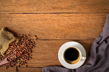 Top view above of Black hot coffee for morning with milk foam for morning menu in white ceramic cup with coffee beans roasted in burlap sack bag on old wood table background. Flat lay with copy space.