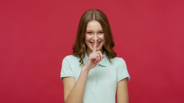 Shh, stop talking! Positive happy young brunette woman in polo t-shirt making quiet gesture, shushing with finger on lips, asking for silence or secrecy. indoor studio shot isolated on red background