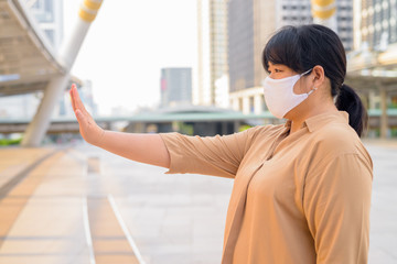 Profile view of overweight Asian woman with mask showing stop gesture in the city