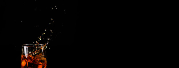An ice cube falls into a glass with whiskey and the spray flies beautifully in different directions on a black background. Panoramic photo for a banner