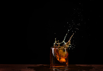 An ice cube falls into a glass with whiskey and splashes fly beautifully in different directions on a black background