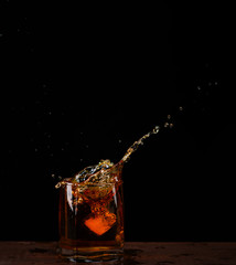 Ice cube falls into a glass with whiskey on a dark background. Spray flying in different directions
