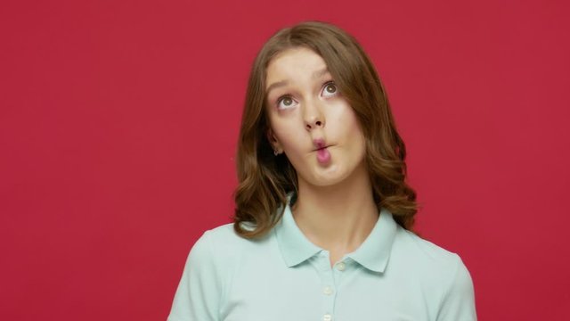 Cute funny young brunette woman in polo t-shirt making wondered comical silly expression with fish face grimace, pecking with pout lips, having fun. indoor studio shot isolated on red background